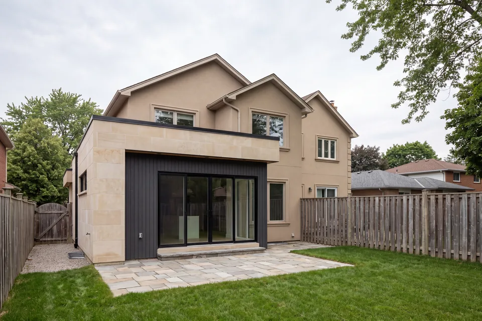 A single-story limestone and dark-siding rear addition with glass doors on an existing two-story house, seen from a fenced backyard